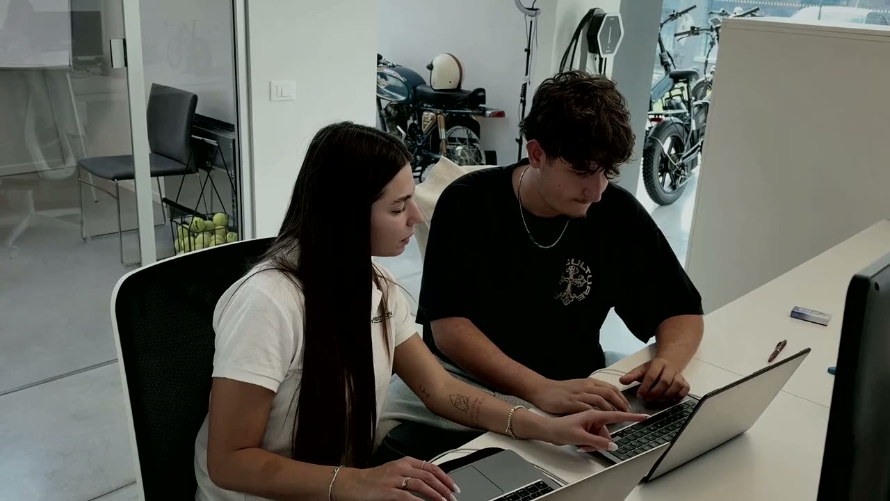 Due persone del team NEXTTOSKIN STUDIO al lavoro sui portatili nello showroom di Modena, con veicoli elettrici sullo sfondo